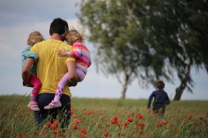 Father of twins walking through a poppy field, photographed from behind. The dark-haired man carries both of his two blond twin daughters on his hips, showing that he is active and committed and has a bond with each of the children.